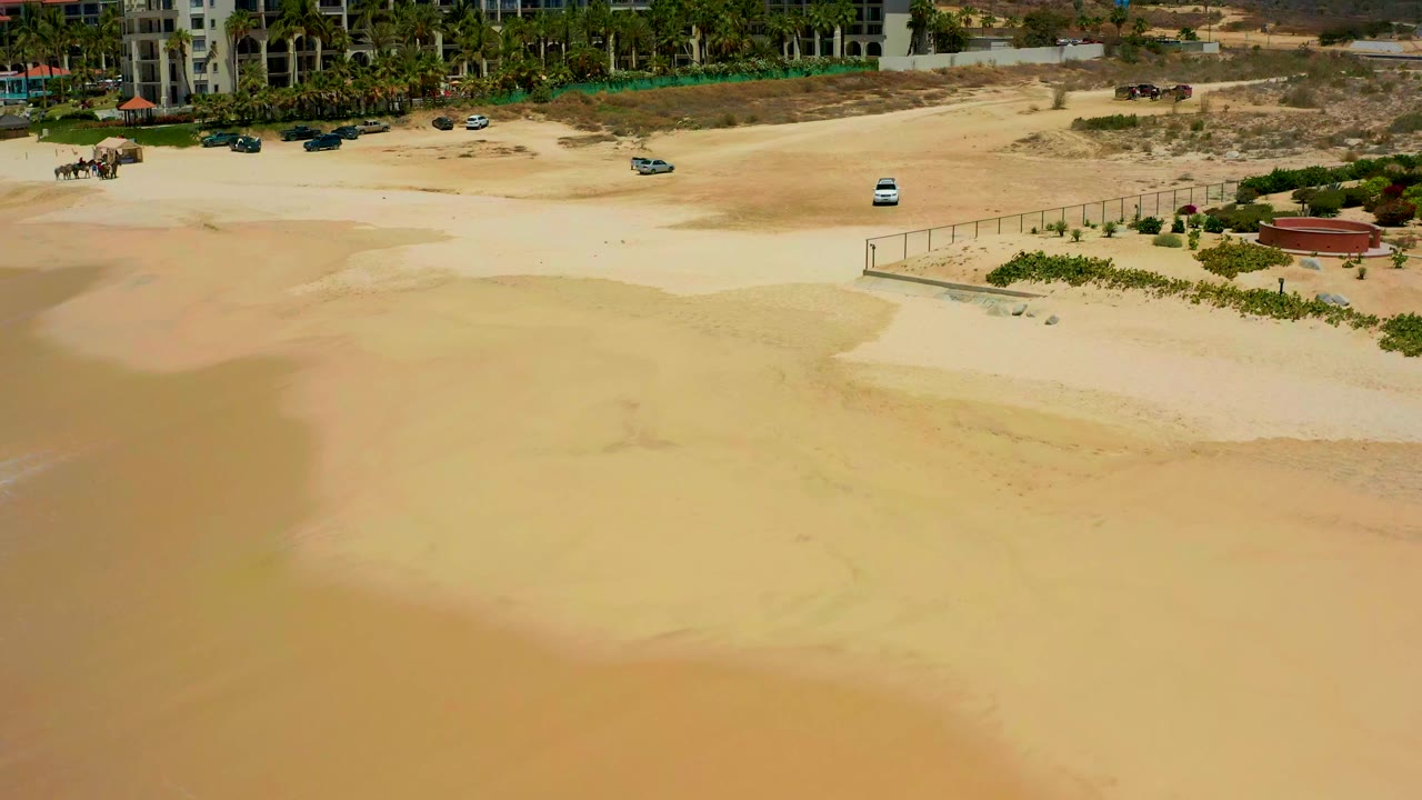 Drone-like aerial frame over Dreams Los Cabos with pale sand below, a fenced edge at right, several parked vehicles near palms, and no visible people.