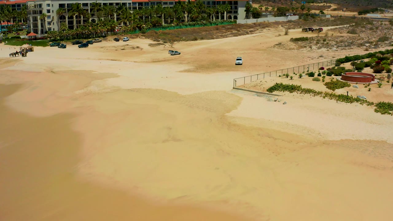 Elevated Dreams Los Cabos view with a bright sand flat in front, fence and landscaped planting to the right, resort buildings behind, and no visible people.