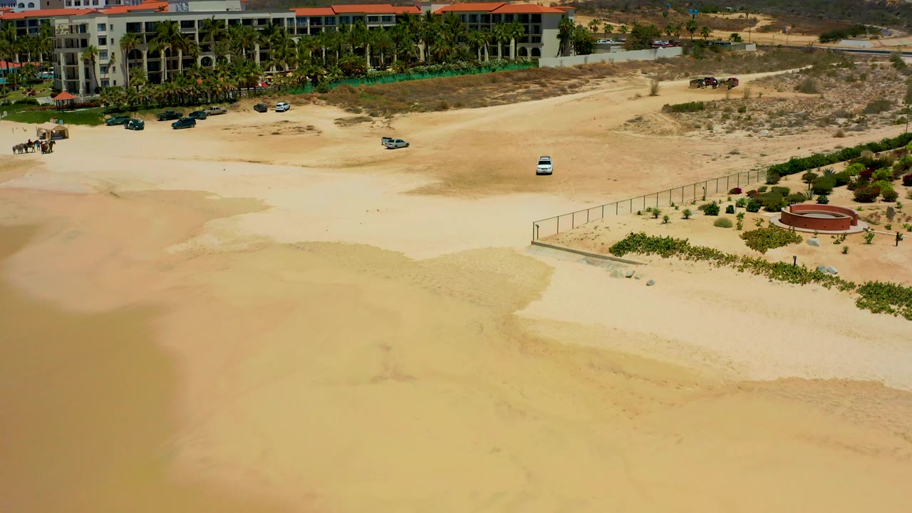Aerial frame at Dreams Los Cabos with wet sand spreading across the lower half, a fenced landscaped corner at right, red-roofed buildings above, and no visible people.