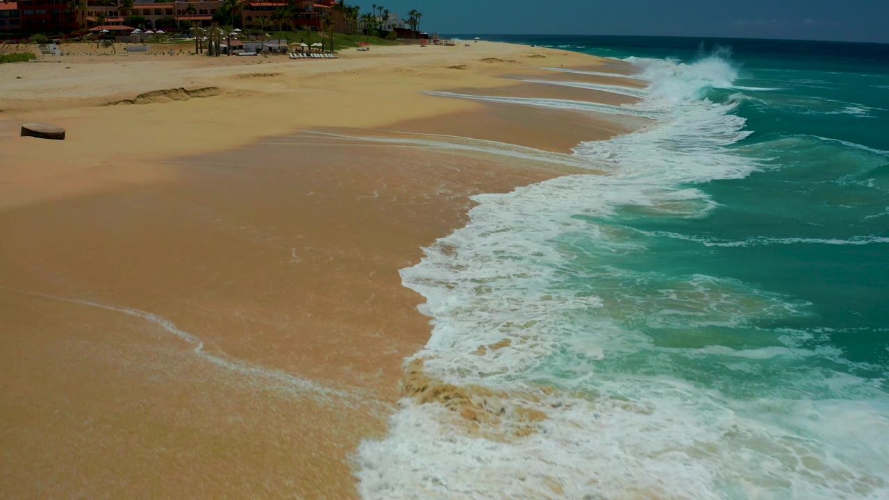 Aerial frame at Dreams Los Cabos showing turquoise surf breaking onto tan sand, a small dark boat on the left beach, resort buildings along the top edge, and no visible people.