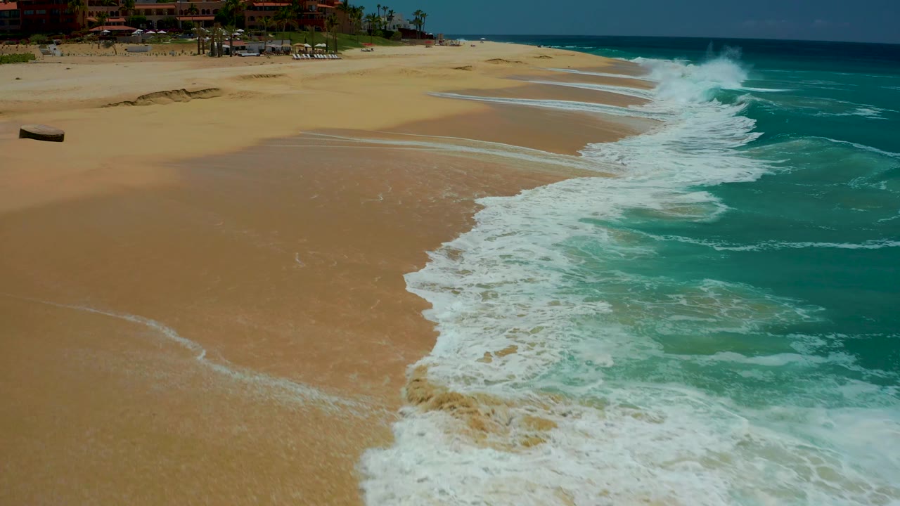 At 0:01, the Dreams Los Cabos shoreline remains in an elevated view, with the small boat near the left edge, white foam pushing farther across wet sand, and no visible people.