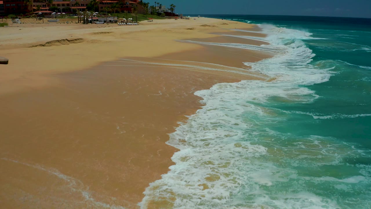 At 0:02, the aerial view shows a broader run of Dreams Los Cabos beach, with the surf line moving along the right side, resort structures at the top, and no visible people.