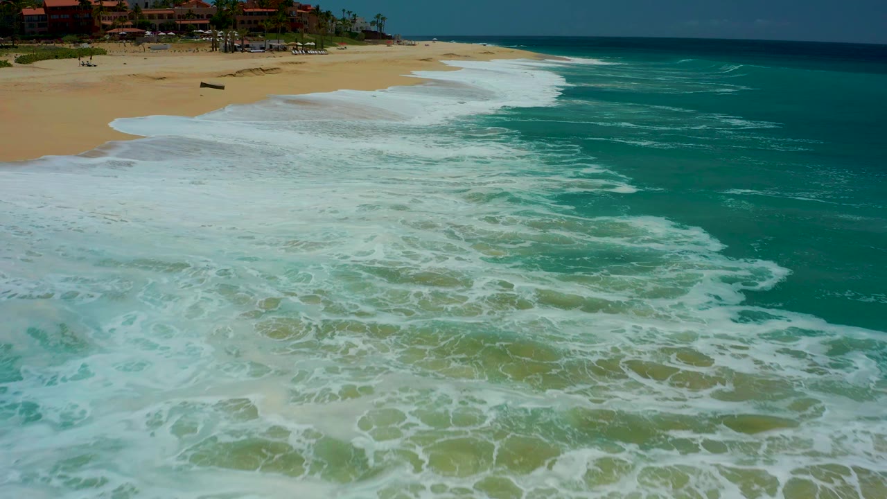 Aerial view of white surf spreading over a sandy beach beside turquoise water.