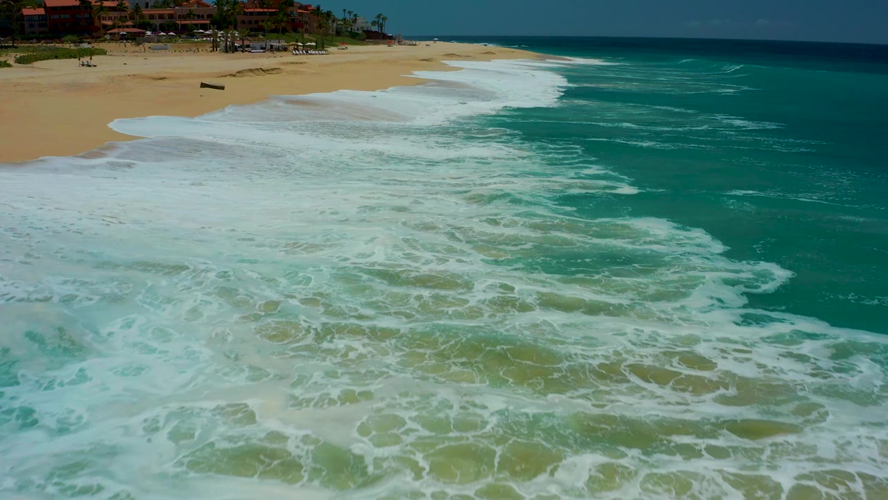 At Dreams Los Cabos in Baja California Sur, the whitewater remains spread across the lower beach while turquoise water curves along the right side and no visible people appear.