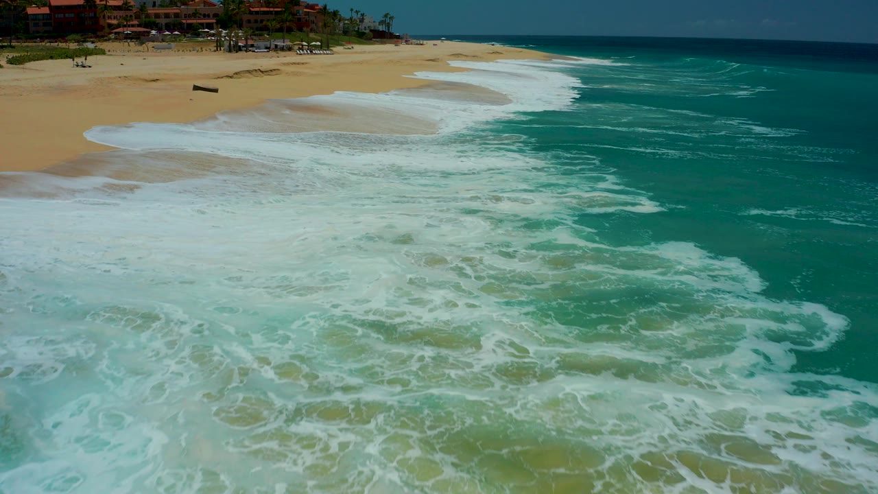 At Dreams Los Cabos in Baja California Sur, the foam line pulls into a long curve across the beach, with more open turquoise water visible on the right and no visible people.