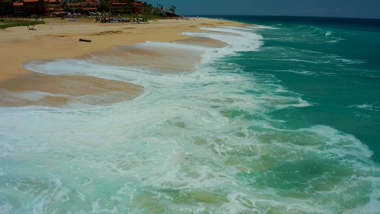 At Dreams Los Cabos in Baja California Sur, receding white foam leaves a broad wet sand pattern while the ocean stays bright turquoise on the right and no visible people are present.