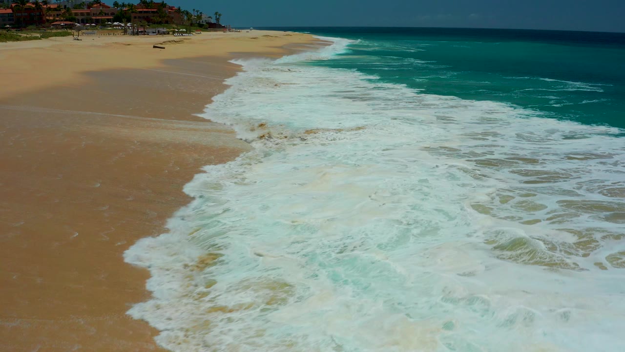At Dreams Los Cabos in Baja California Sur, white surf pushes diagonally over tan sand with turquoise ocean to the right and no visible people.