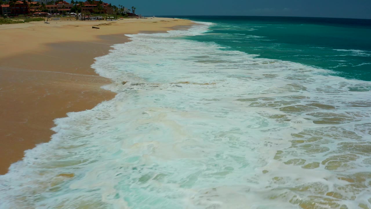 At Dreams Los Cabos in Baja California Sur, broad white foam fills the foreground, the turquoise ocean sits beyond the break, and the upper beach remains empty with no visible people.