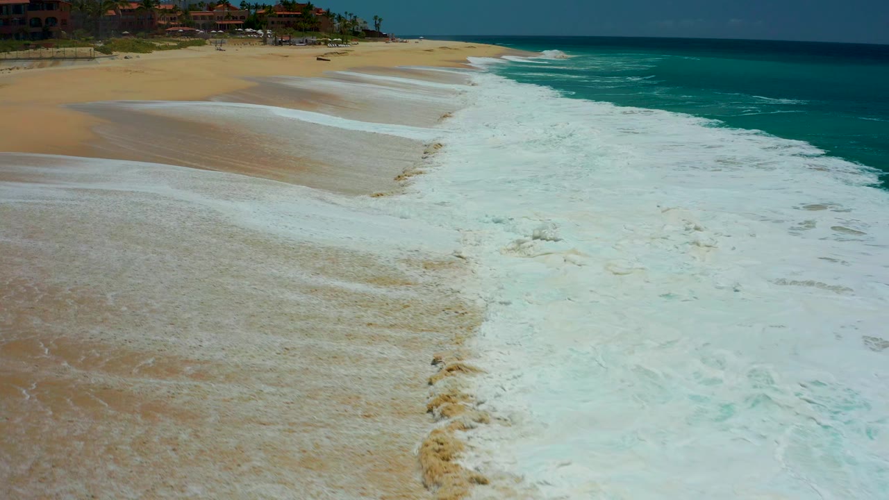 Aerial view of white surf washing over tan beach beside turquoise ocean.