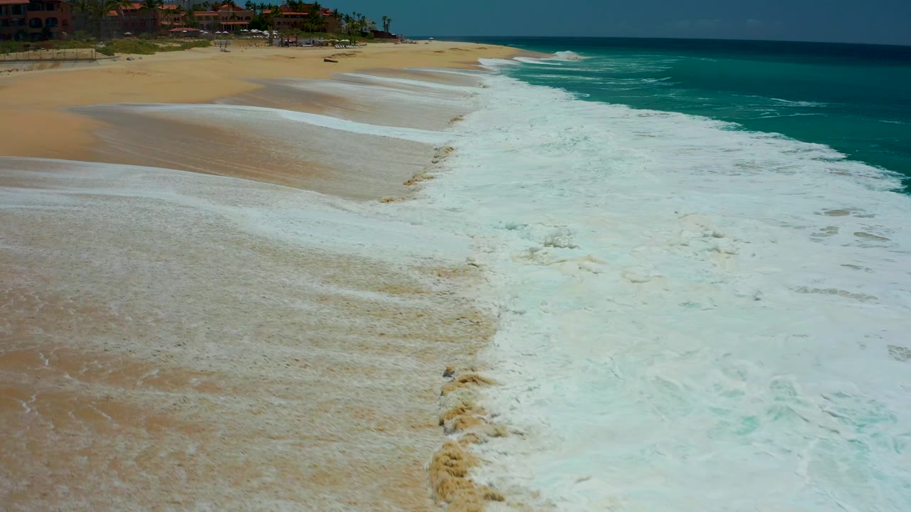 Whitewater fills the center of the Dreams Los Cabos shoreline as wet tan sand remains visible at left and blue-green ocean extends along the right edge, with no visible people.
