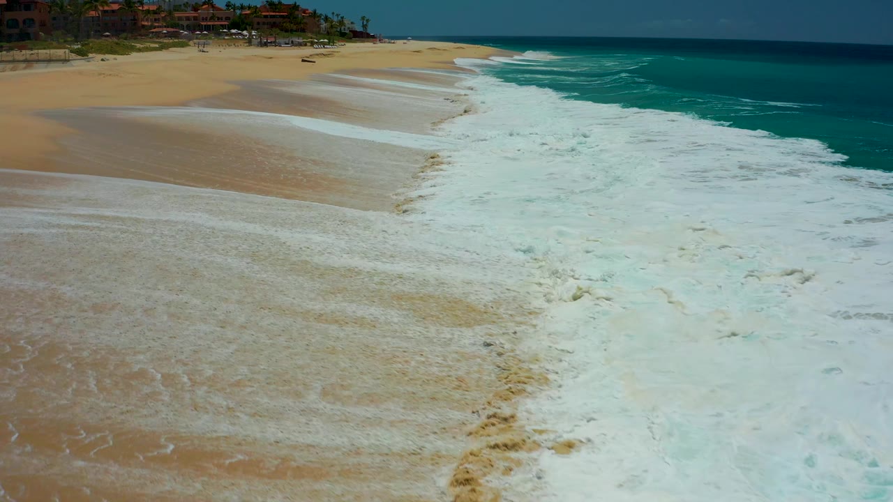 The surf line at Dreams Los Cabos pulls diagonally across the frame, leaving more exposed wet sand on the left while darker turquoise water holds the right side, with no visible people.