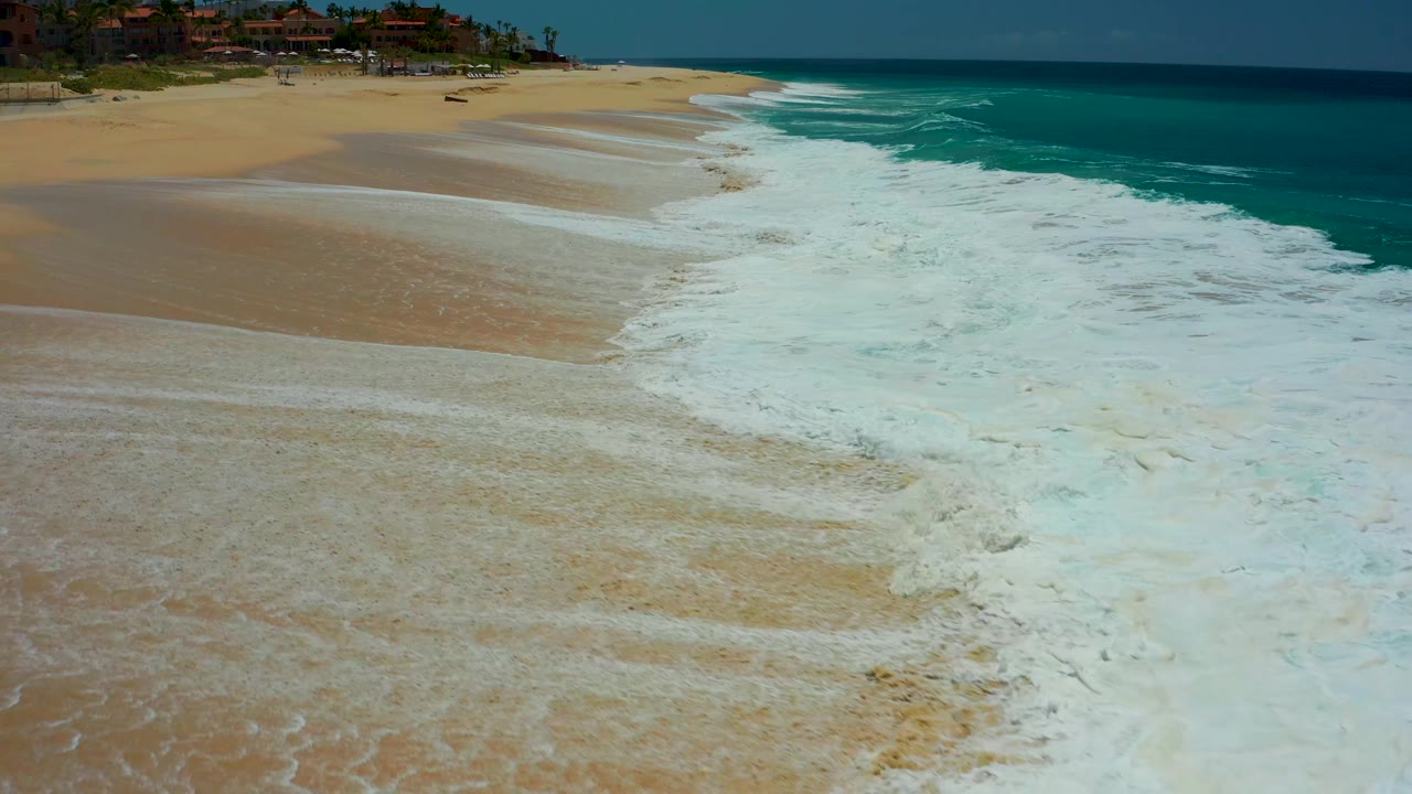 White surf occupies the lower and right portions of the Dreams Los Cabos beach while a smooth tan sand section opens at left and resort structures remain small in the background, with no visible people.