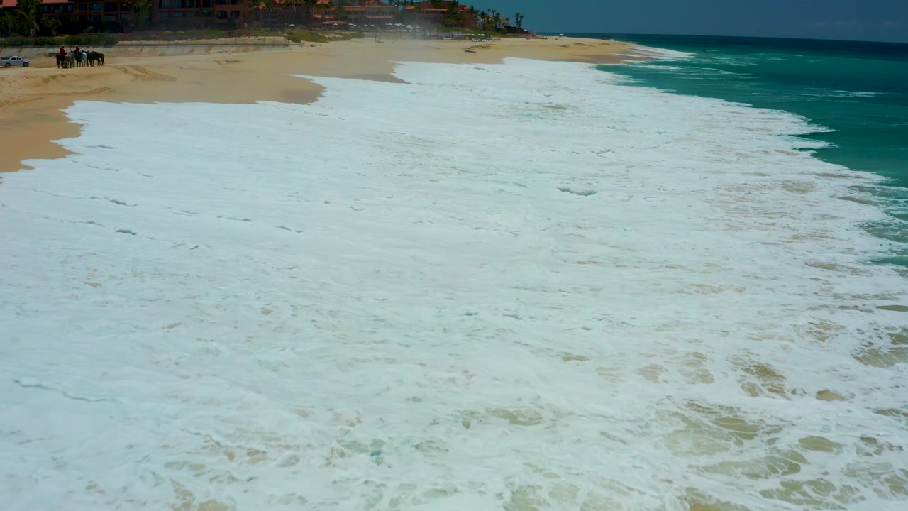 At Dreams Los Cabos in Baja California Sur, foamy white surf covers most of the pale sand while turquoise water runs along the right side, with no visible people.