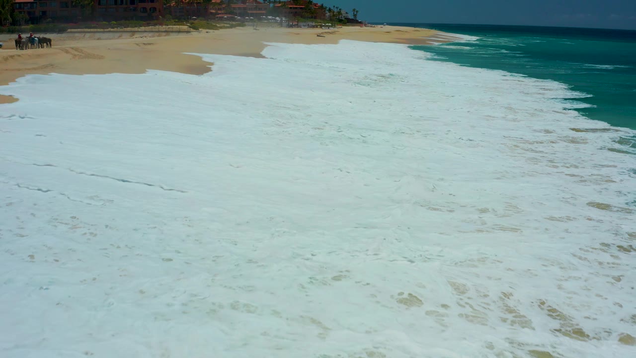 At Dreams Los Cabos in Baja California Sur, a broad sheet of white surf foam blankets the beach while the turquoise sea remains visible along the right edge, with no visible people.