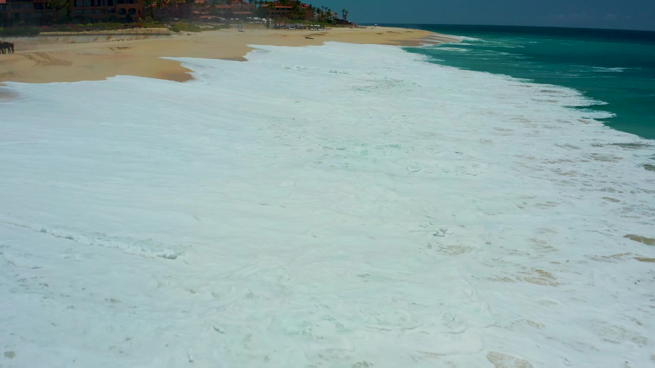 At Dreams Los Cabos in Baja California Sur, foamy water spreads across the foreground sand as a diagonal shoreline separates pale beach from turquoise ocean, with no visible people.
