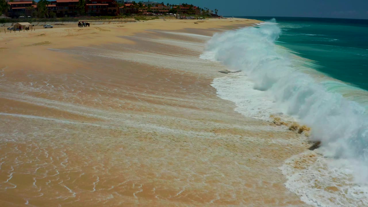 At Dreams Los Cabos in Baja California Sur, the wave has advanced across the beach, leaving a broad white foam edge over wet tan sand with the ocean still visible to the right.