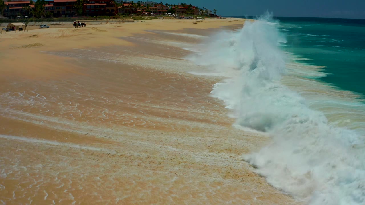 At Dreams Los Cabos in Baja California Sur, the wave throws a higher burst of white spray near the right side of the beach while thin foam streaks mark the wet sand.
