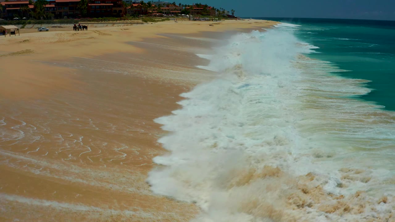 At Dreams Los Cabos in Baja California Sur, a thick diagonal band of white surf rolls over the beach, with tan sand still exposed on the left and blue-green ocean on the right.