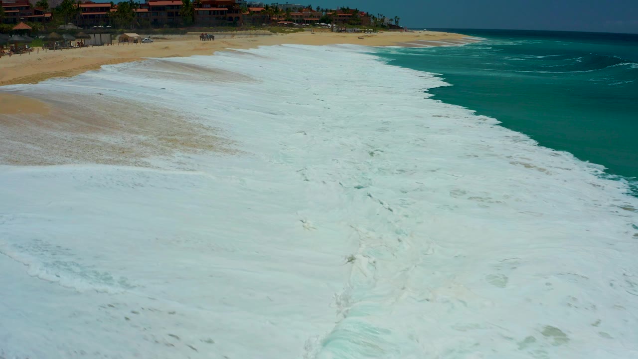 At Dreams Los Cabos in Baja California Sur, white foam covers much of the wet sand while turquoise water sits to the right and distant beachfront buildings line the top edge, with no visible people.