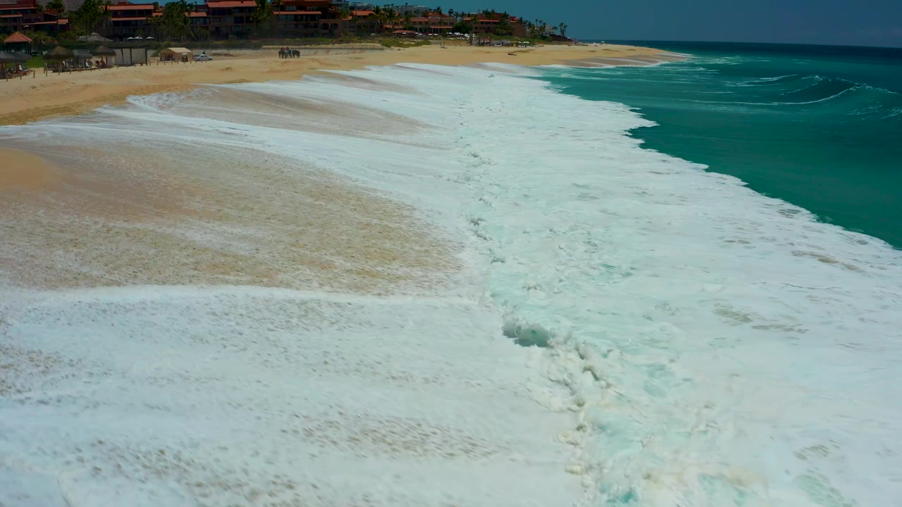The foam edge pulls back from the upper sand while a turquoise wave line remains offshore at Dreams Los Cabos, leaving streaks of whitewater across the wet beach and no visible people.