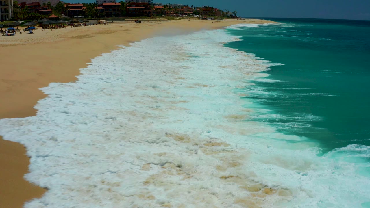 Aerial view at Dreams Los Cabos in Baja California Sur showing white foam covering the lower beach, turquoise ocean on the right, coastal buildings and palms at the top, and no visible people.