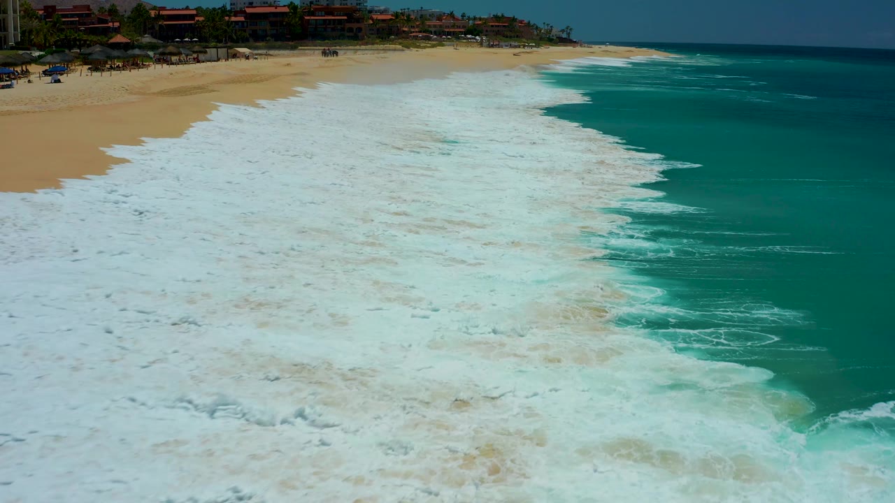 Aerial view over Dreams Los Cabos surf where white foam covers much of the sandy shore, darker turquoise water sits to the right, resort-area rooftops line the top, and no visible people appear.