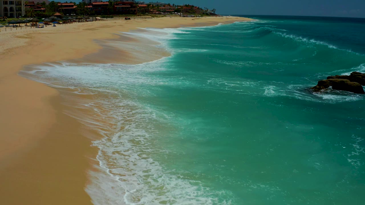 The surf line advances across the light brown sand at Dreams Los Cabos, with turquoise water to the right, resort-area buildings in the distance, and no visible people.