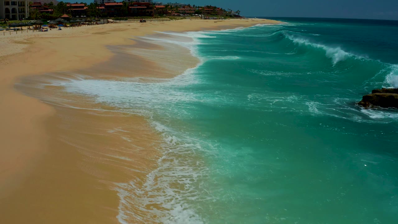 A larger turquoise wave curls near the right edge while white wash reaches the sand at Dreams Los Cabos, with distant beachfront structures and no visible people.