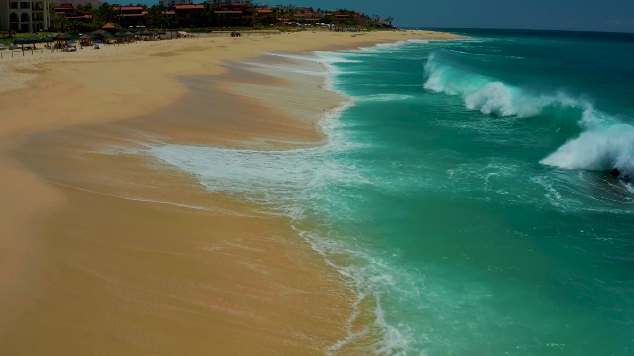 Whitewater builds from the right side of the frame and pushes toward the open sand at Dreams Los Cabos, with the turquoise ocean behind it and no visible people.