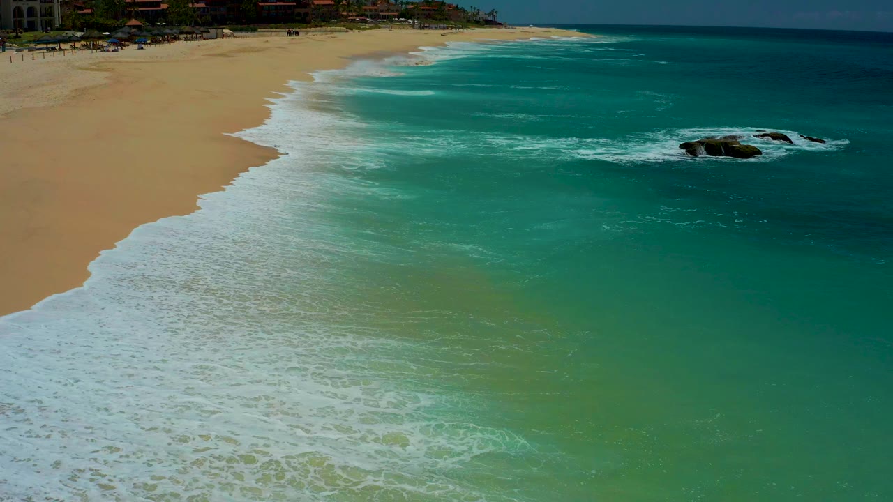 Aerial frame from Dreams Los Cabos showing turquoise water and white surf spreading across sand, with dark rocks offshore at right and no visible people.