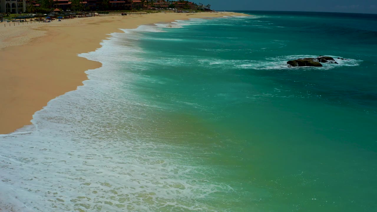 At 0:02, foamy waves spread wider over the Dreams Los Cabos sand while the nearshore water appears green-blue and the offshore rocks sit near the right edge, with no visible people.