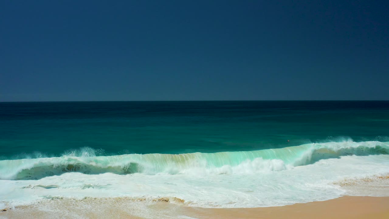At Dreams Los Cabos in Baja California Sur, blue ocean water rolls toward a sandy shore with white foam across the lower frame and no visible people.