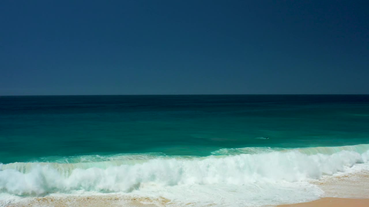 White surf surges across the foreground while deep blue ocean and turquoise shallows fill the frame at Dreams Los Cabos, with no visible people.
