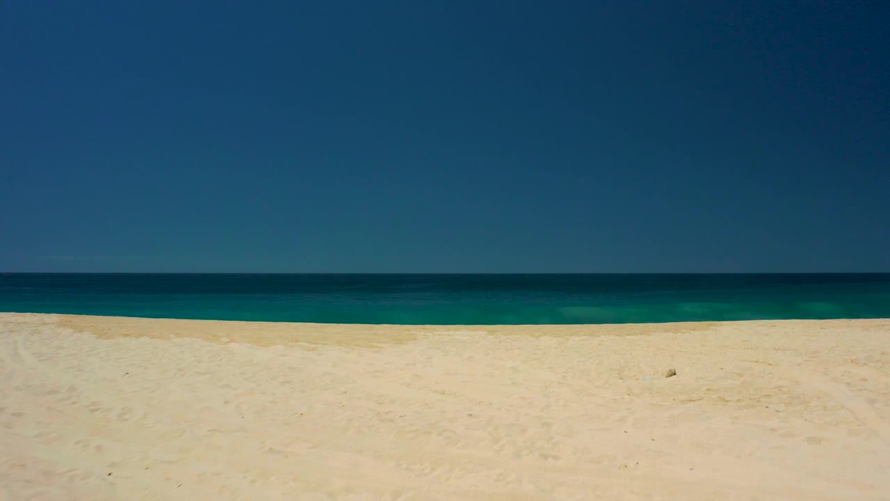 Empty sandy shoreline at Dreams Los Cabos with turquoise water near shore, darker ocean beyond, and no visible people.