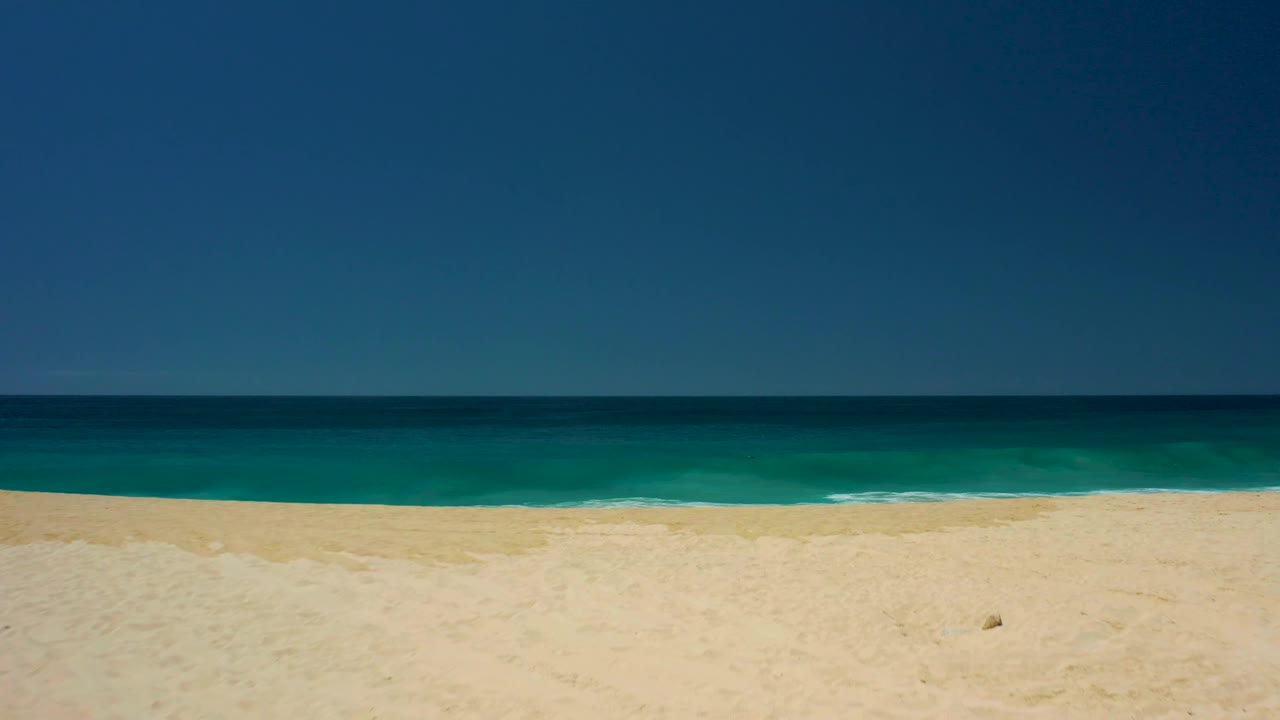 Sandy shore at Dreams Los Cabos with a narrow turquoise band, deep blue open water, clear sky, and no visible people.