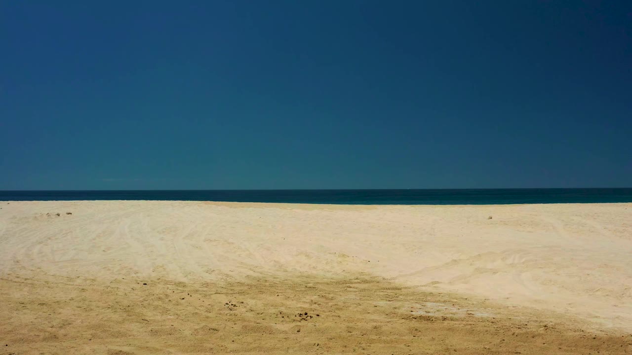 At 0:01, the Dreams Los Cabos shoreline remains empty, with textured sand and faint track marks below a narrow dark blue ocean band and clear sky, with no visible people.