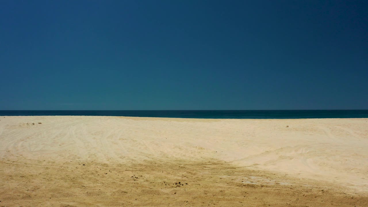 At 0:03, the beach remains wide and empty at Dreams Los Cabos, with a dark blue ocean strip crossing the background and smooth clear sky above, with no visible people.