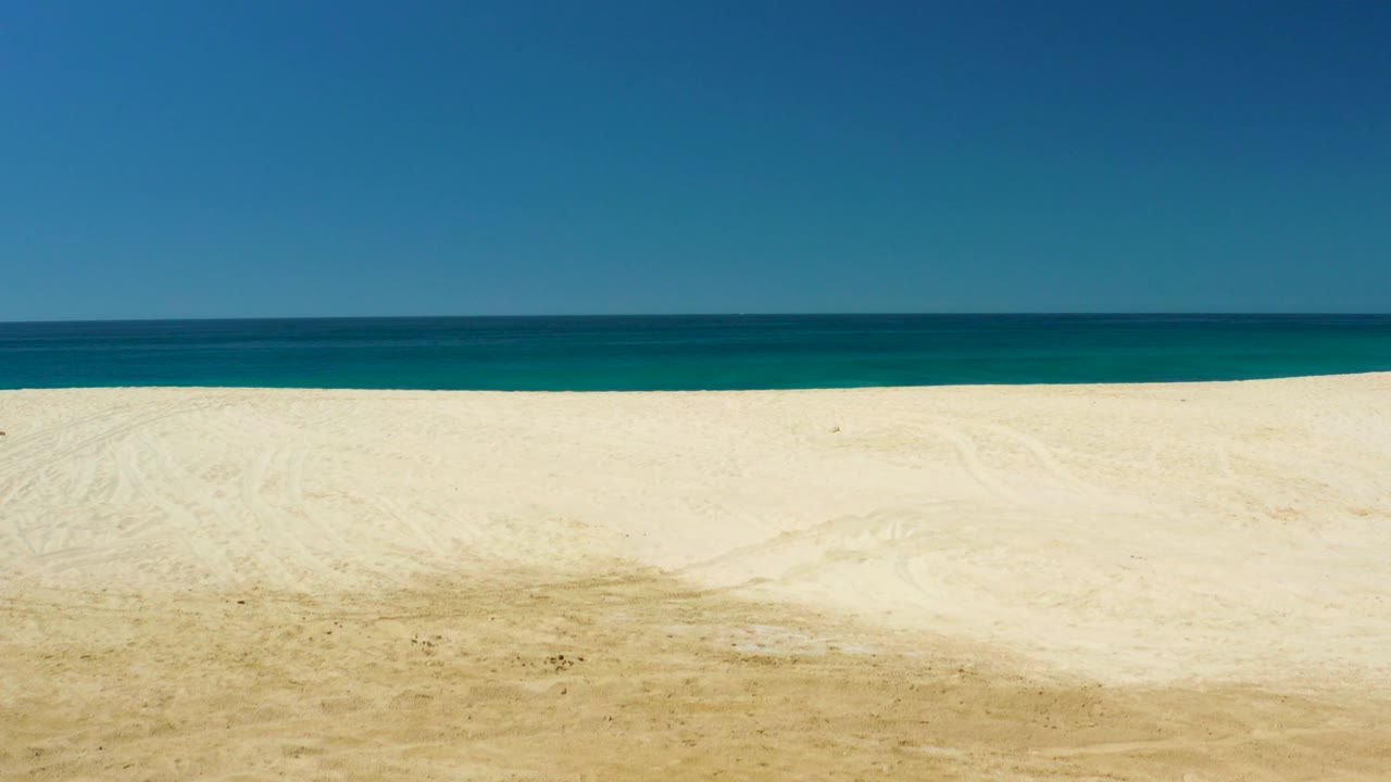 The frame shows the same open Dreams Los Cabos shoreline with pale sand, a low horizontal ocean strip, uninterrupted blue sky, and no visible people or vehicles.