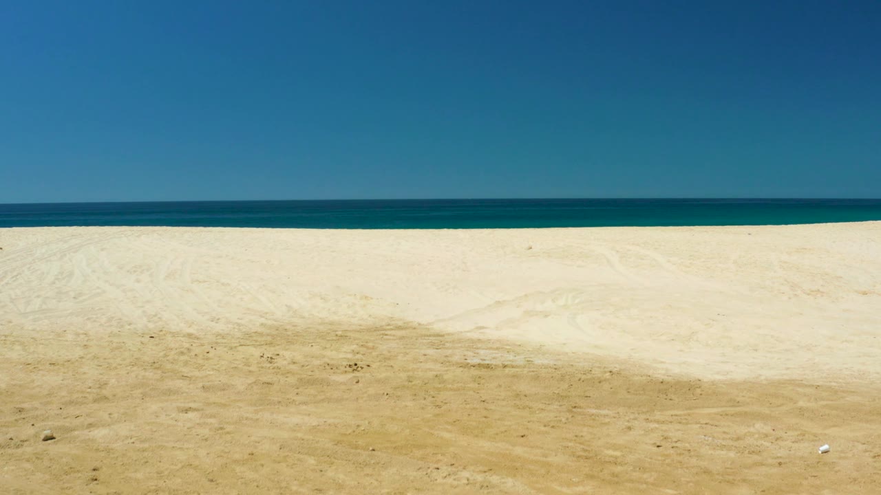 This frame shows an empty sandy shore at Dreams Los Cabos, with faint tire-like textures in the sand, a flat ocean horizon, clear blue sky, and no visible people.
