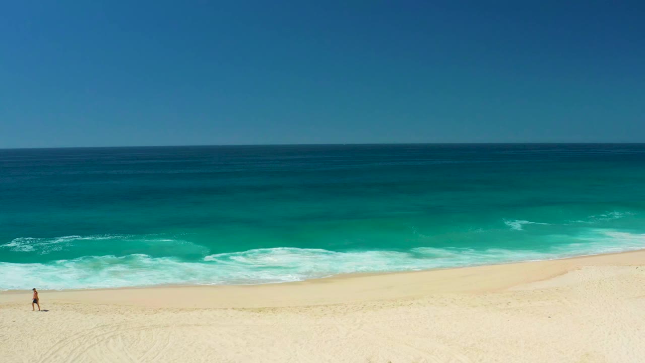 At Dreams Los Cabos in Baja California Sur, Mexico, a distant person appears near the left edge of a wide sandy beach while turquoise waves break under a clear sky.