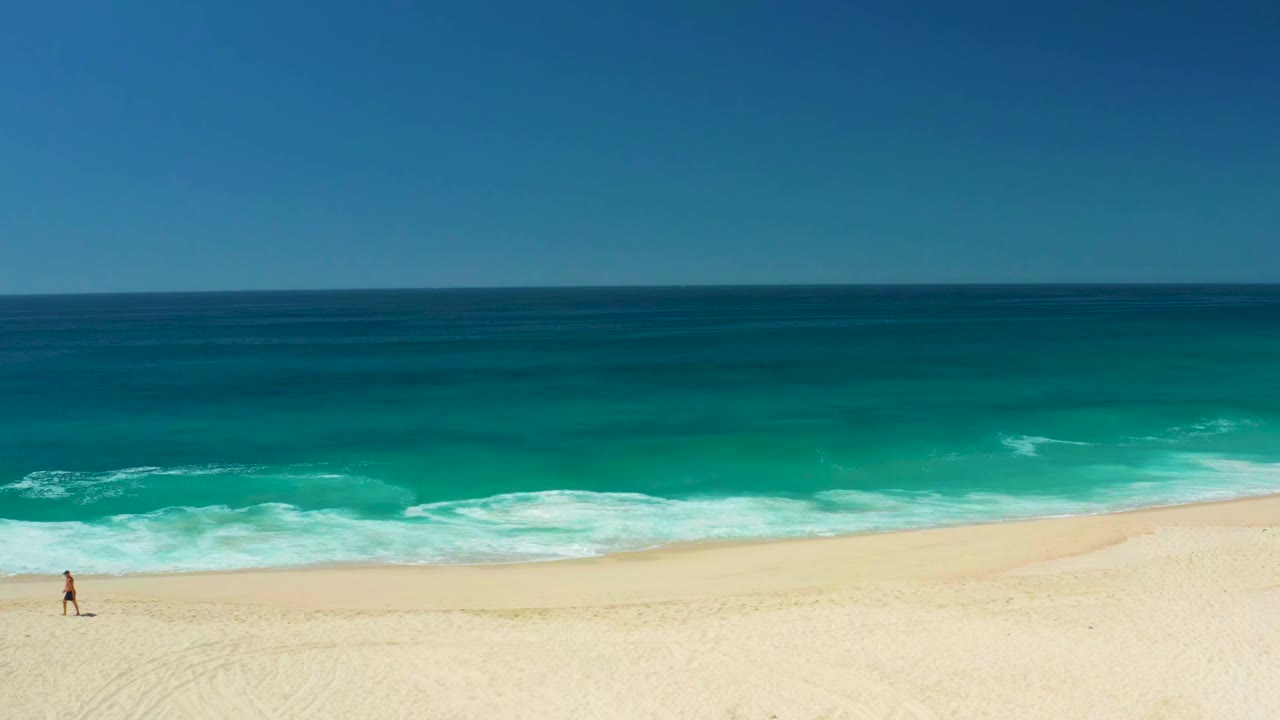 Small person walking across pale sand with ocean surf behind them.