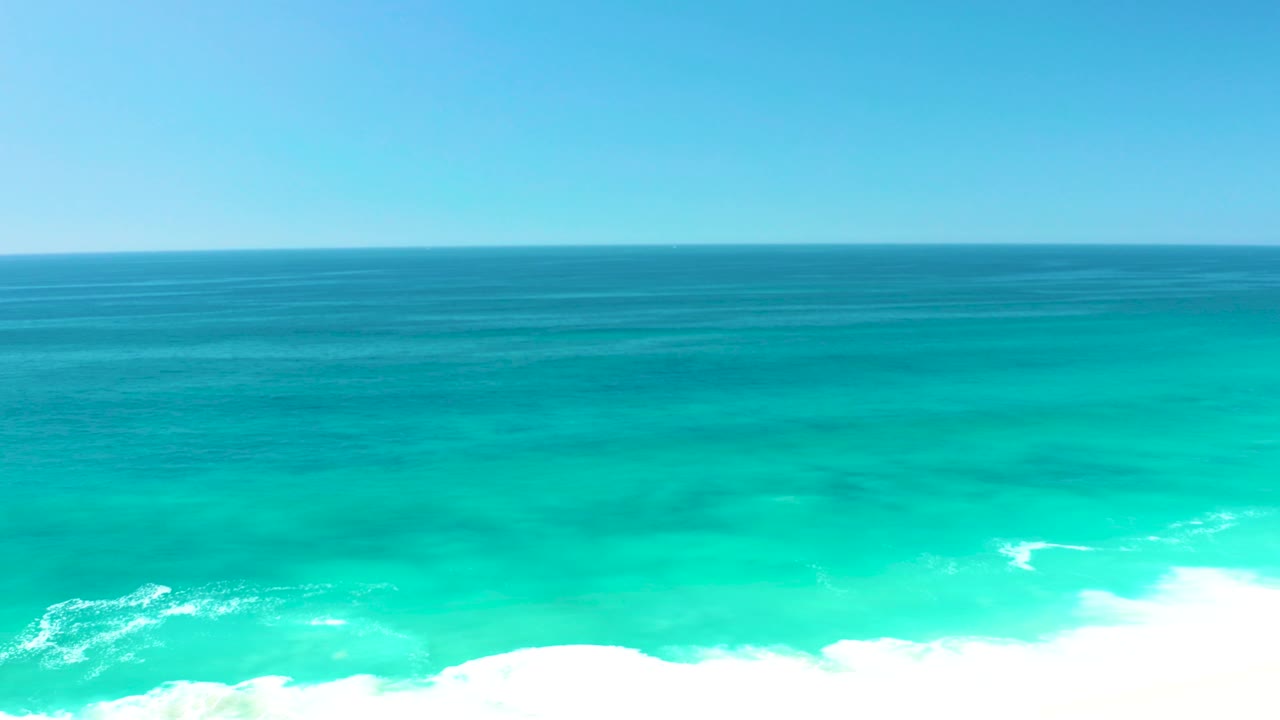A wide ocean gradient runs from darker blue near the horizon to bright turquoise near shore, with white wave foam clustered along the lower edge and no visible people.