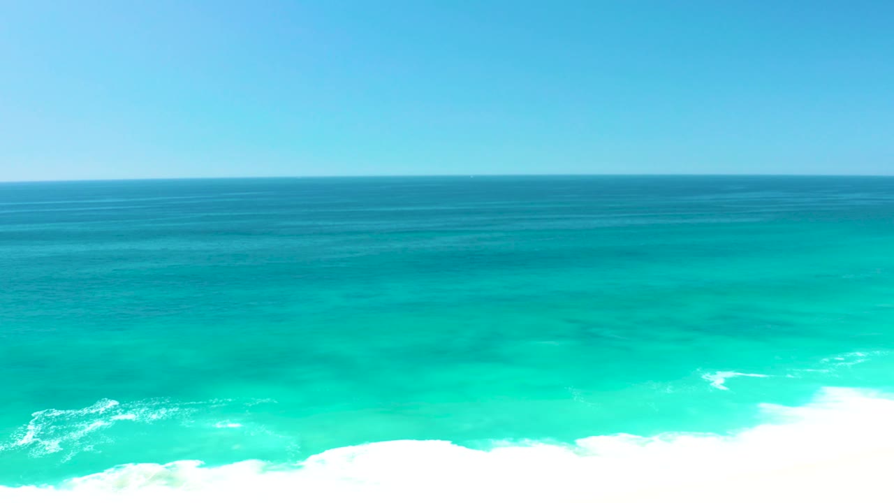 Clear sky sits above a flat ocean horizon while turquoise coastal water and a white breaking wave occupy the lower portion, with no visible people.