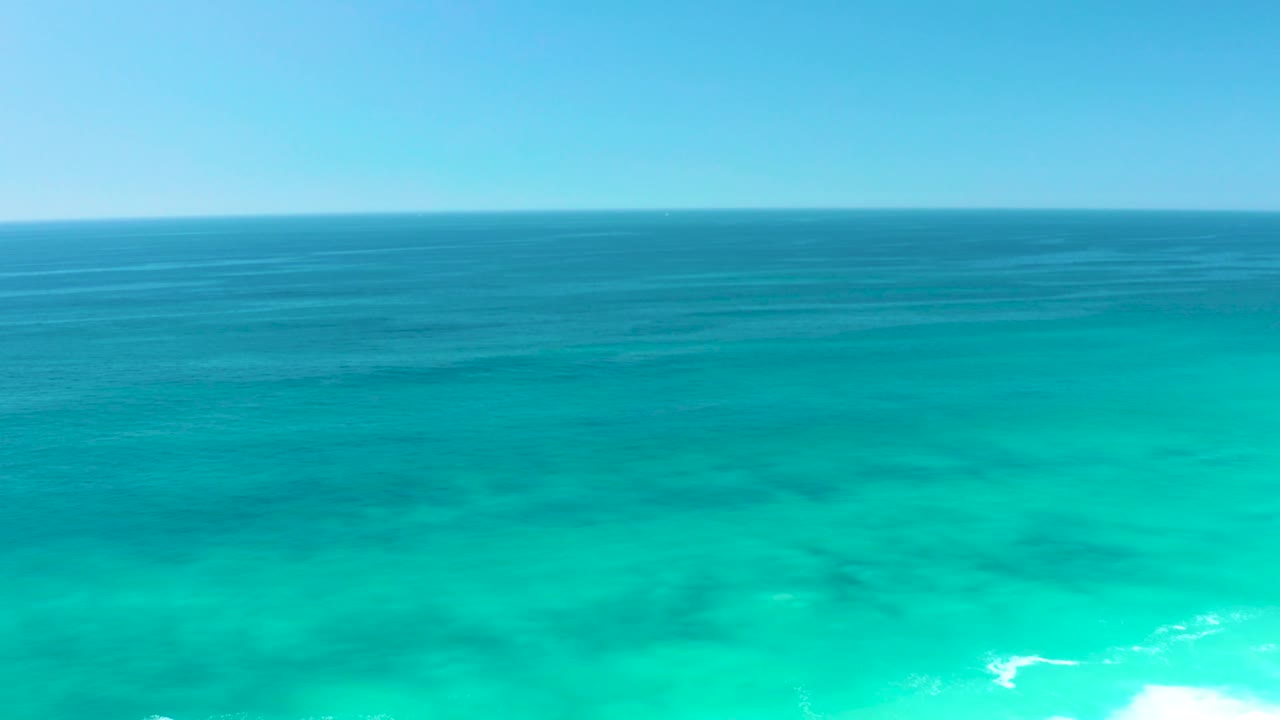 At Dreams Los Cabos, turquoise water stretches below the horizon while a small patch of white surf begins to show near the lower right, with no visible people.