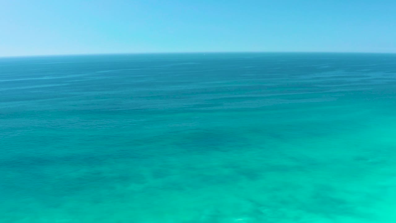 Elevated view of clear turquoise shallows at Dreams Los Cabos, with darker blue water bands stretching toward a level horizon and no visible people.