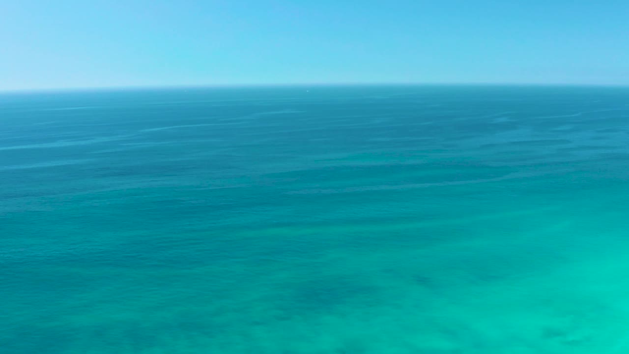 Frame at 0:02 from Dreams Los Cabos, with the distant ocean line, layered blue water, green-turquoise shallows, subtle wave streaks, and no visible people.