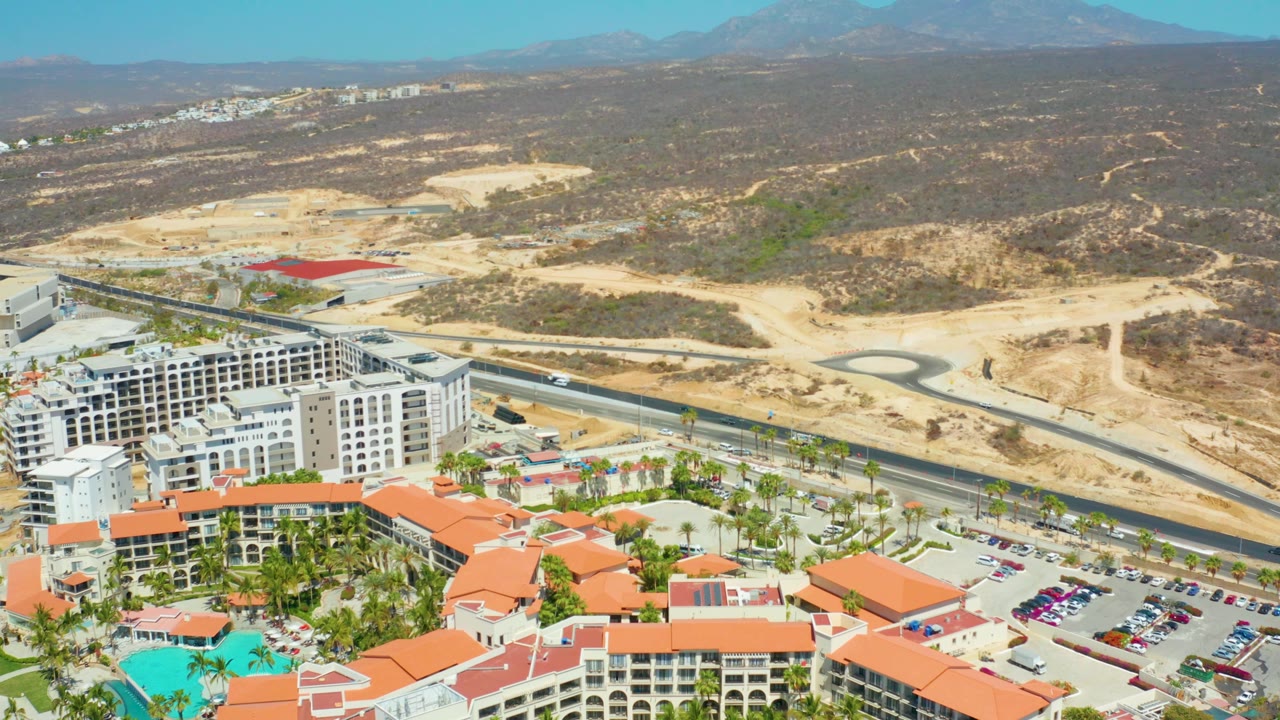 Second frame over Dreams Los Cabos with resort roofs and pool at the bottom, parked cars and a road through the middle, dry hillside terrain beyond, and no visible people.