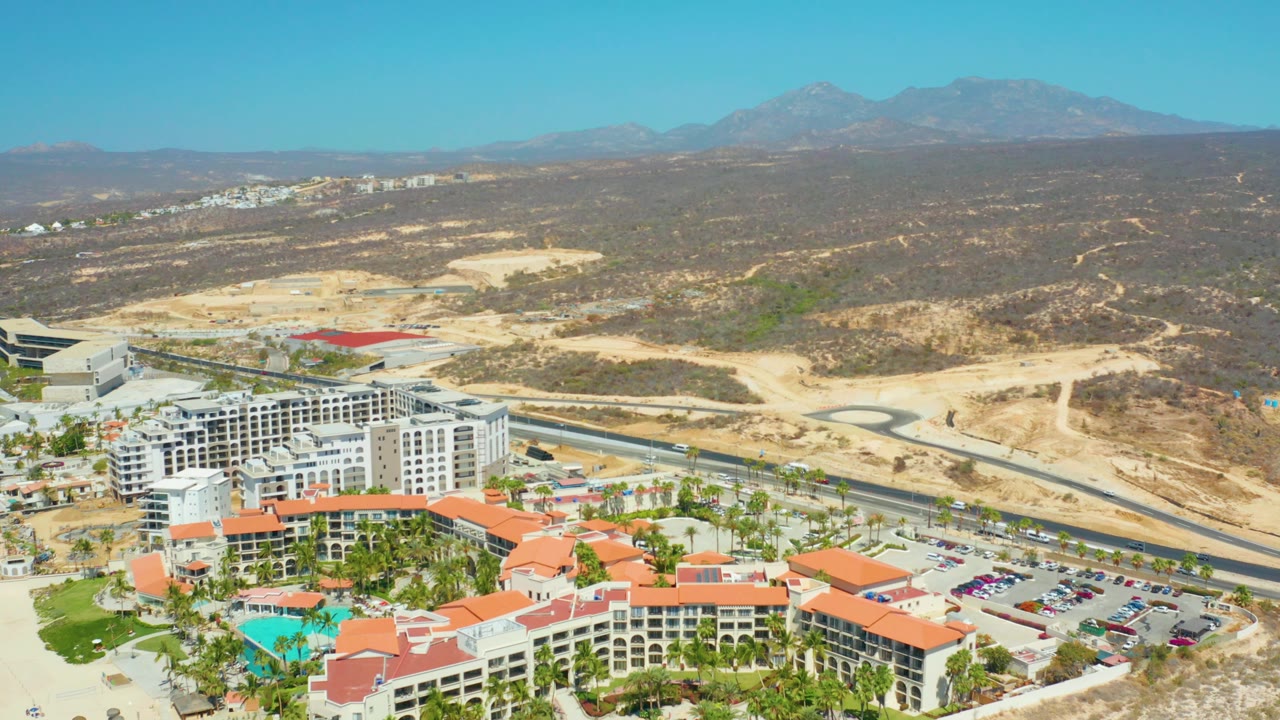 Opening aerial frame over Dreams Los Cabos shows red-tiled resort roofs, a pool, parking lots, a nearby road, dry hills, distant mountains, and no visible people.