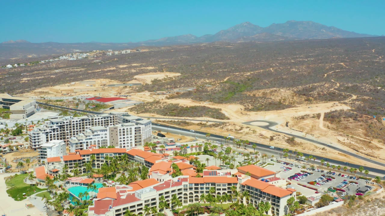 At two seconds, Dreams Los Cabos remains in the lower frame with red roofs, parking areas, and palm trees, while a road cuts past dry hills toward distant mountains, with no visible people.