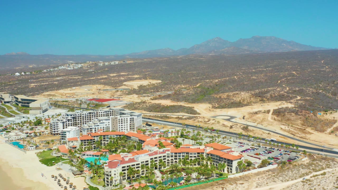 Aerial view at 0:01 over Dreams Los Cabos, showing more of the resort's white buildings and red roofs beside pools, beach sand, a road, dry terrain, mountains, and no visible people.